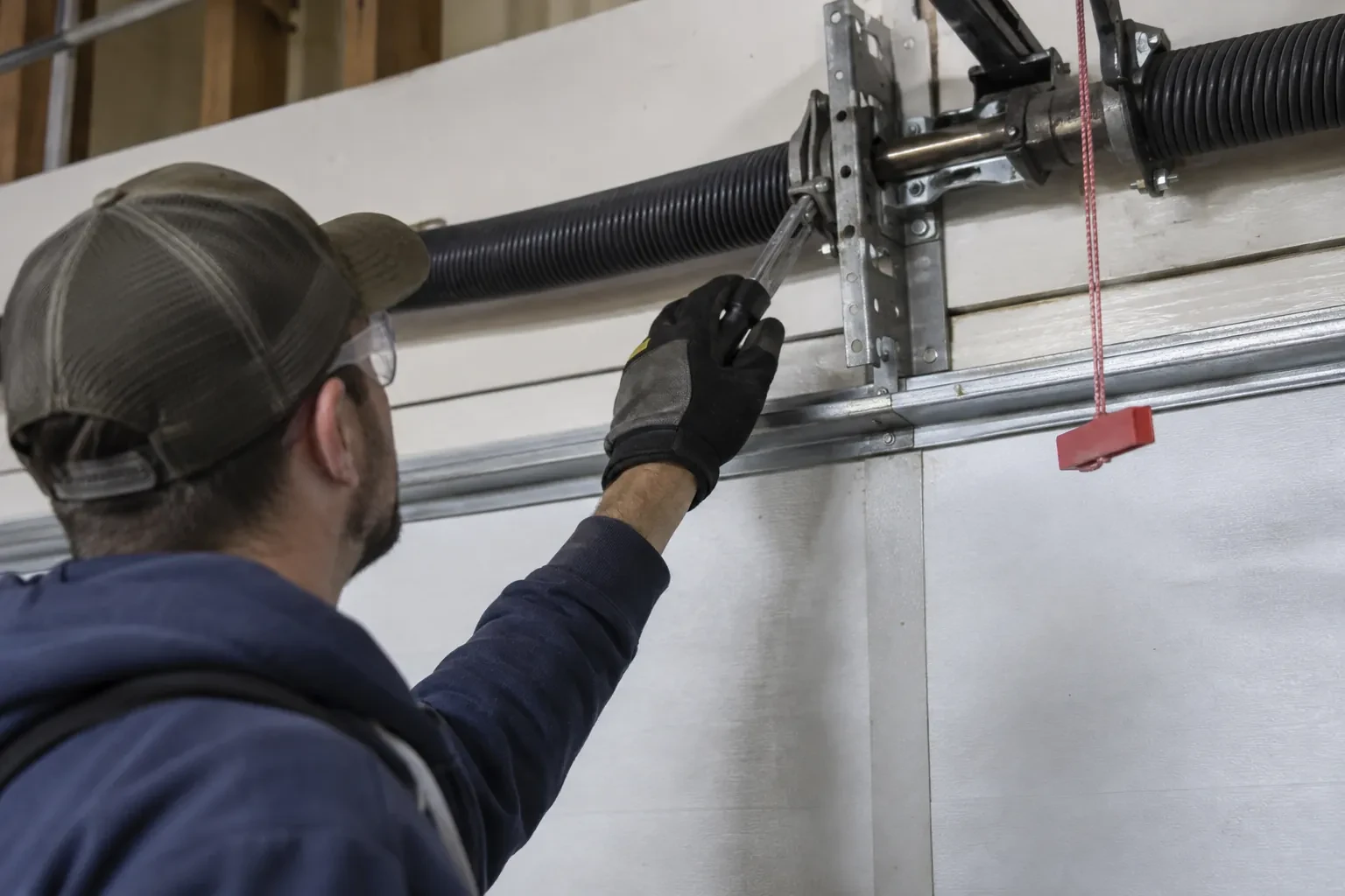 Technician in a cap reaching a gloved hand with a tool toward a coiled spring and bracket above a garage door. — Ashburnham