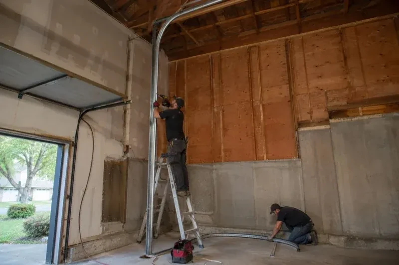 One person on a stepladder working beside a tall curved garage-door track while another kneels beside loose track sections in an unfinished garage. — High-Lift Garage Door Conversion, Peterborough