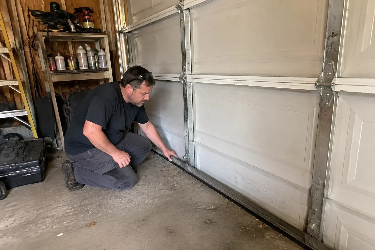 Person kneeling by a white paneled door in a garage, one hand on the black seal along its bottom edge. — Garage Door Roller Replacement, Peterborough